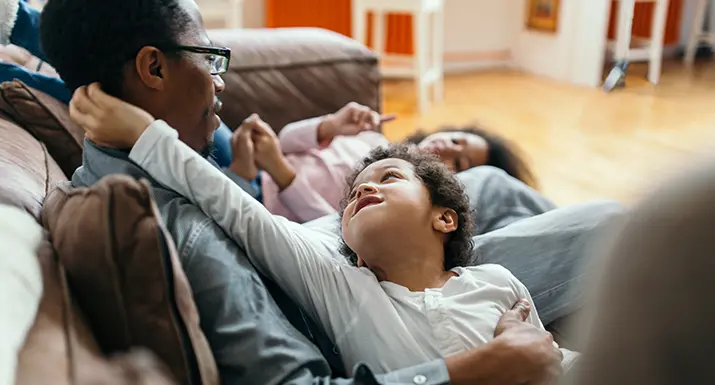 Människor ligger på en brun soffa inomhus. Ett barn tittar uppåt med armarna runt en vuxen.